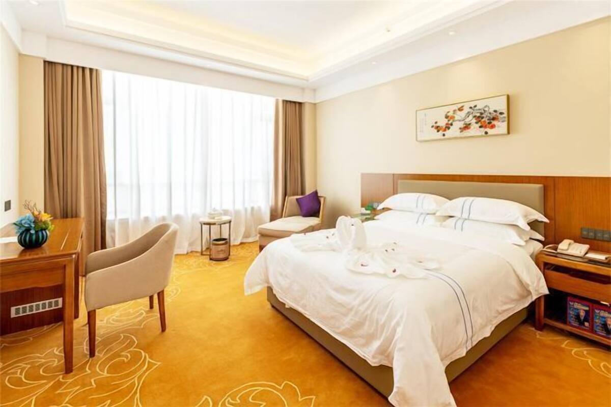 Quarto de hotel com uma cama grande, uma escrivaninha com cadeira, uma mesa pequena e uma mesa de cabeceira.