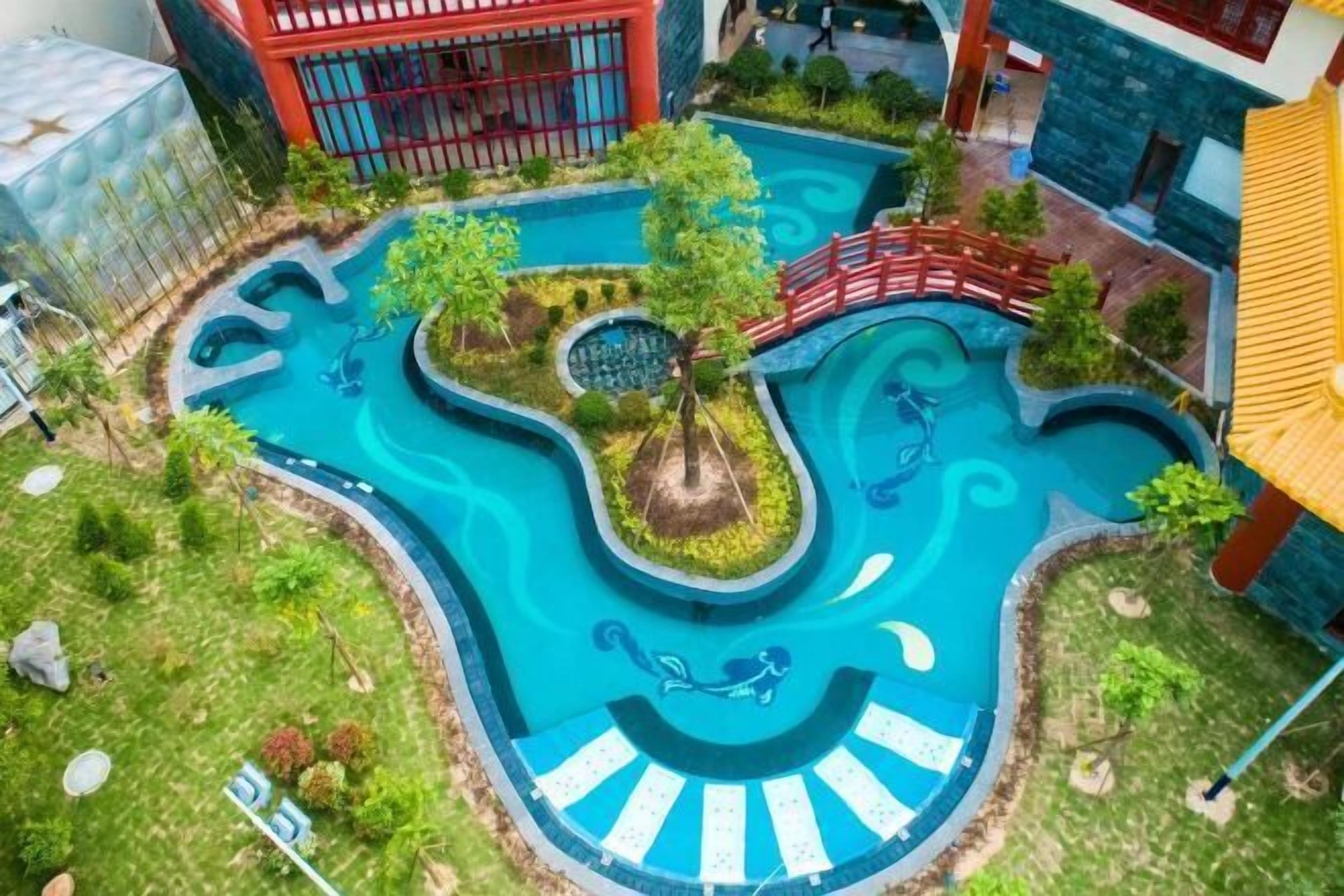 Vue aérienne d’un complexe de piscines comprenant plusieurs bassins reliés entre eux, entouré de verdure et surplombé par un pont rouge.