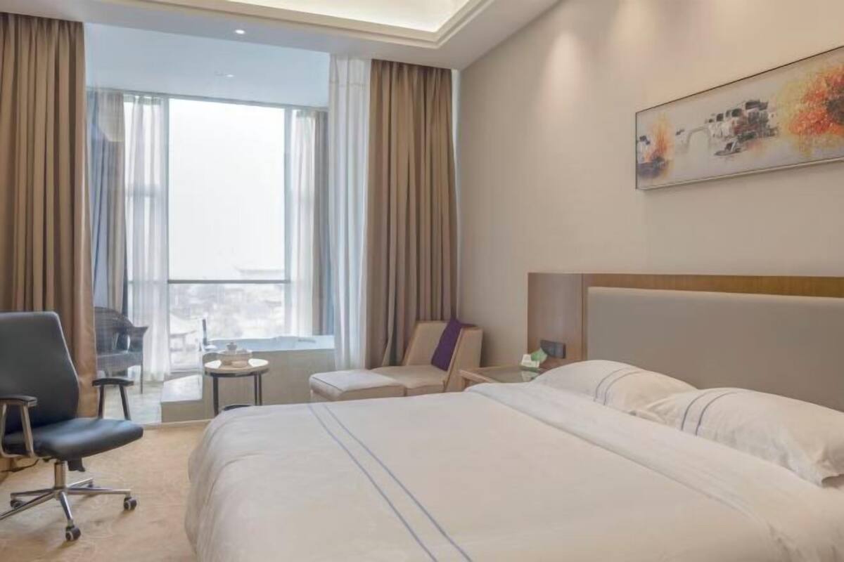 Quarto de hotel com uma cama grande, uma escrivaninha, uma cadeira e vista da cidade através de amplas janelas.