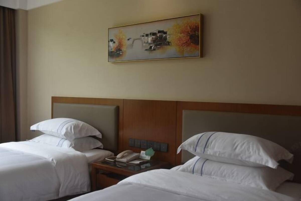 Quarto de hotel com duas camas, cabeceira de madeira, mesa de cabeceira com telefone e caixa de lenços, e um quadro na parede.