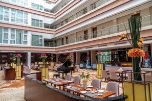 A multi-level hotel lobby with a grand piano, dining tables, and a central fountain.