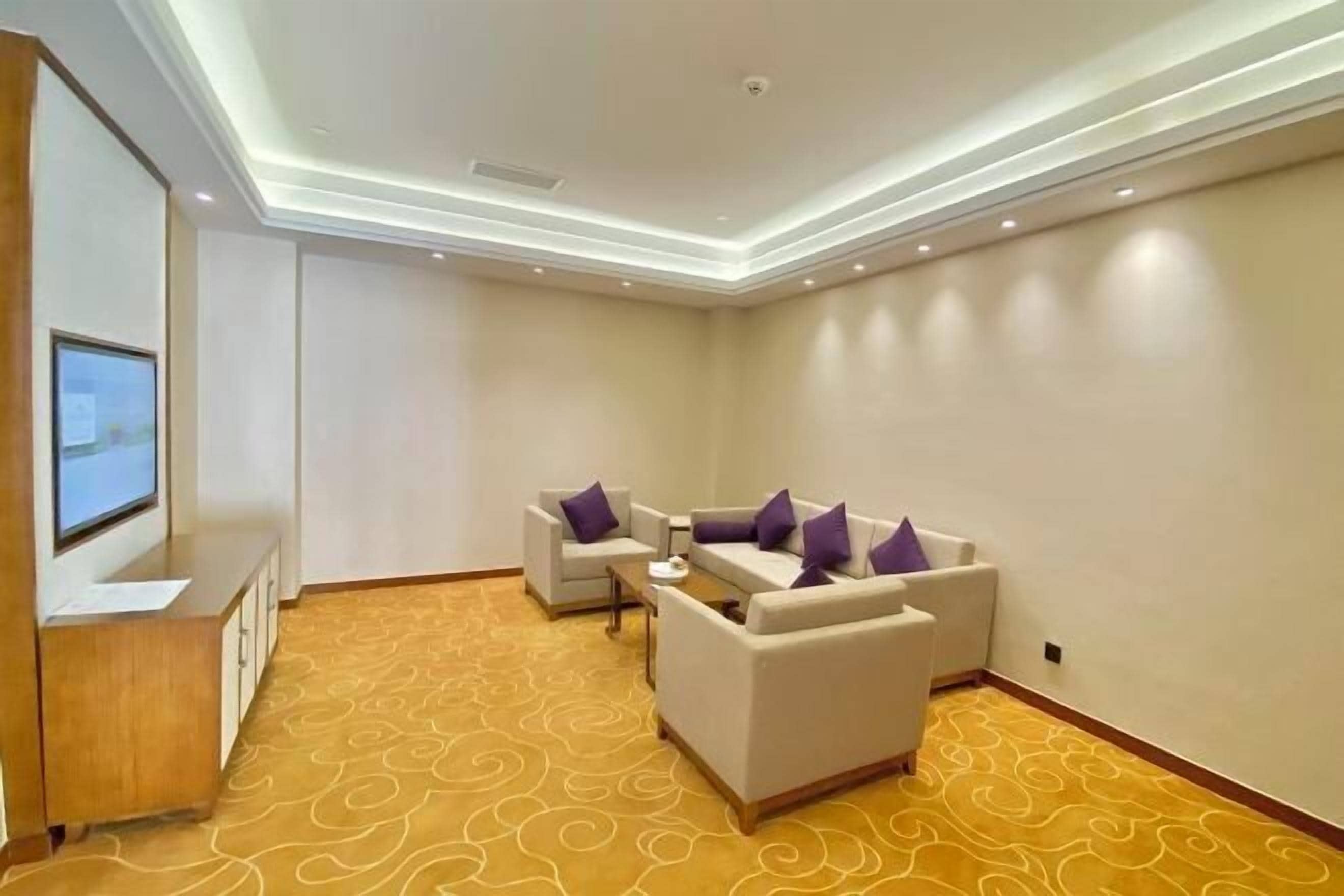 A hotel room with a seating area featuring two sofas and a coffee table, a flat-screen TV mounted on a wooden cabinet, and a patterned carpet.