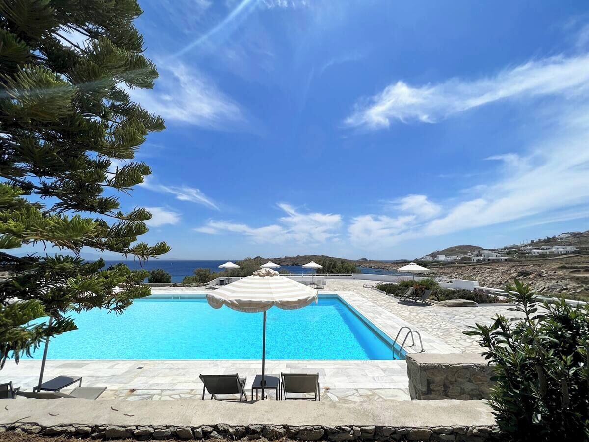 A swimming pool with lounge chairs and umbrellas, surrounded by stone tiles and greenery, with a view of the sea and hills in the background.