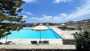 Una piscina con sillones de descanso y sombrillas, rodeada de baldosas de piedra y vegetación, con vista al mar y colinas al fondo.