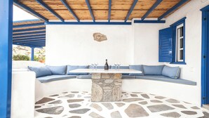 Un patio con suelo de piedra, un banco integrado, una mesa, contraventanas azules y un muro de piedra.