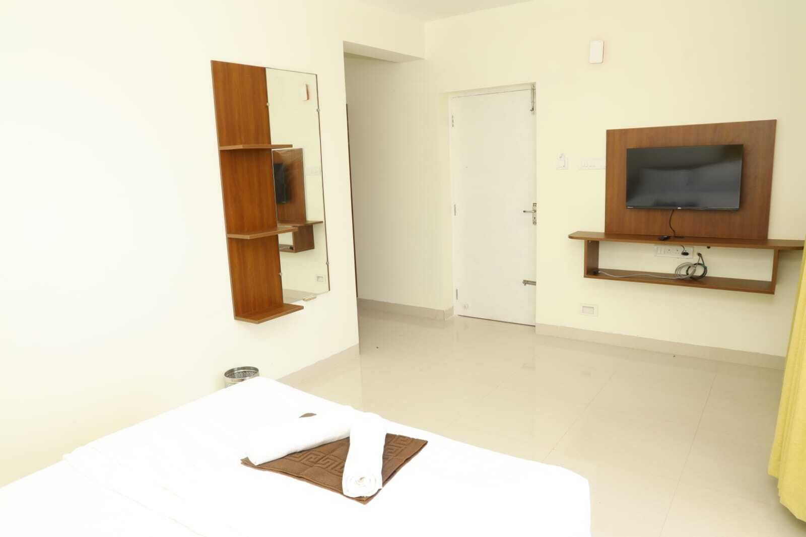 A hotel room with a white bed, a wooden shelf with a mirror, a television on a wooden stand, and a white door.