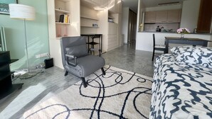 A modern hotel room with a grey armchair, a patterned rug, a bed with a blue and white duvet, a small desk, and a kitchenette in the background.