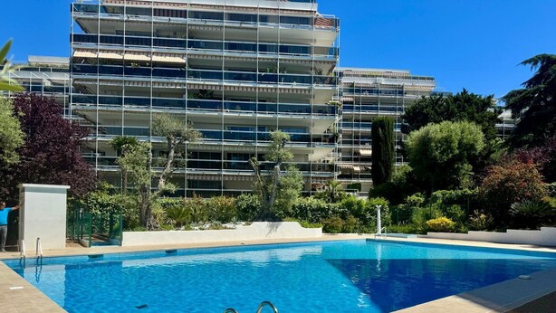 A swimming pool with a clear blue water, surrounded by a tiled deck. In the background, there is a multi-story building with balconies and greenery.