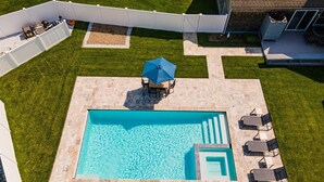 Piscina retangular com deck revestido, cercada por uma grade branca, e área de varanda com mesa e cadeiras.