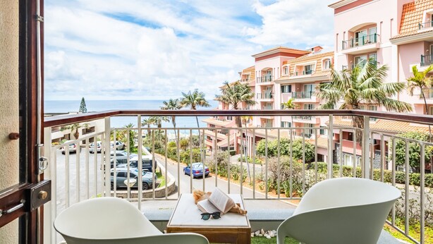 A balcony with two white chairs, a small table with a book, sunglasses, and a hat. The balcony overlooks a street with parked cars and a building with pink and orange facades. - Summertime Stay by Homie (Funchal)