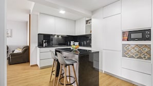 A modern kitchen with white cabinetry, a black countertop, and a built-in microwave. There are two bar stools and a small island with a fruit bowl. - Summertime Stay by Homie (Funchal)