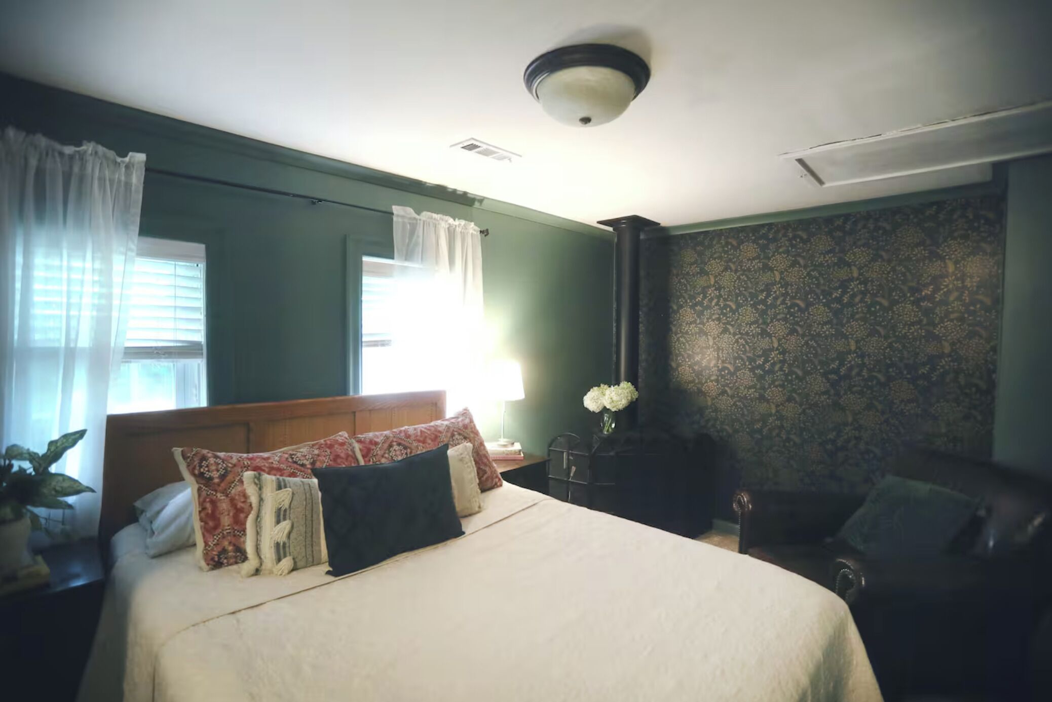 Un dormitorio con cama, mesita de noche, silla, ventana con cortinas y papel tapiz estampado en la pared.