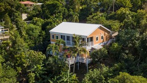 Une maison à deux étages, entourée d’une végétation tropicale luxuriante, avec un balcon bien visible et un toit blanc.