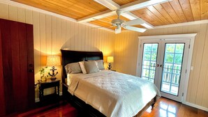 Une chambre avec un plafond en bois, un lit recouvert d’une couette blanche, deux lampes de chevet et une vue à travers une porte vitrée.