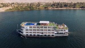 Un gran barco crucero fluvial con varias cubiertas y una piscina, amarrado a un río con un paisaje desértico al fondo.