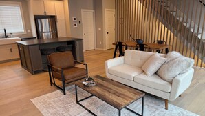 A modern living room with a sofa, coffee table, and a chair. There is a kitchen area with a refrigerator and cabinets. A staircase is visible in the background.
