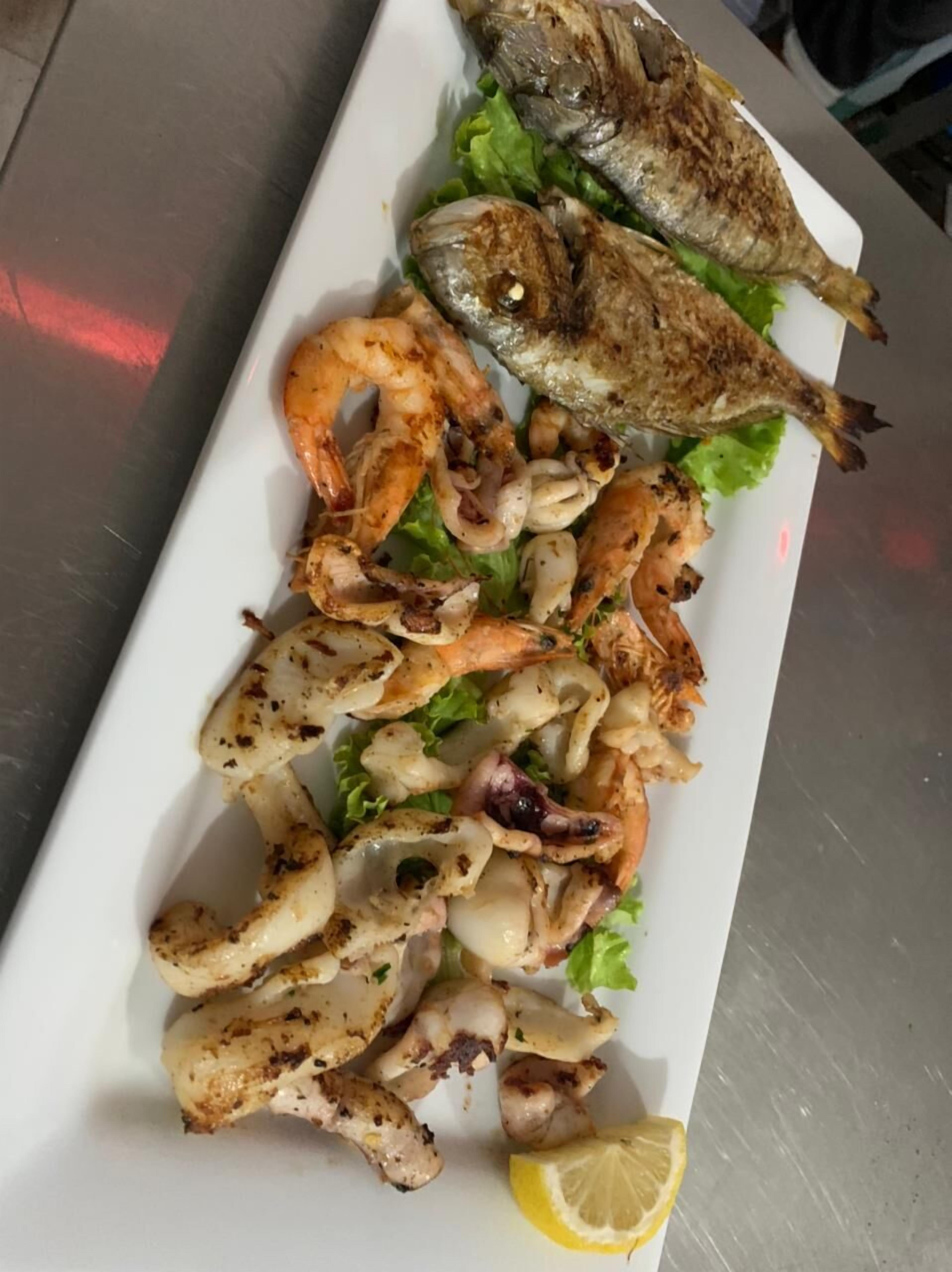 Un plato rectangular blanco con pescado a la parrilla, camarones y calamares, adornado con lechuga y un gajo de limón.