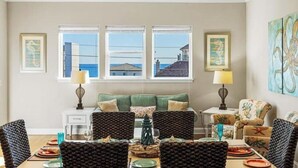 Ein Essbereich mit einem grünen Sofa, gemusterten Stühlen und Blick auf das Meer durch große Fenster.