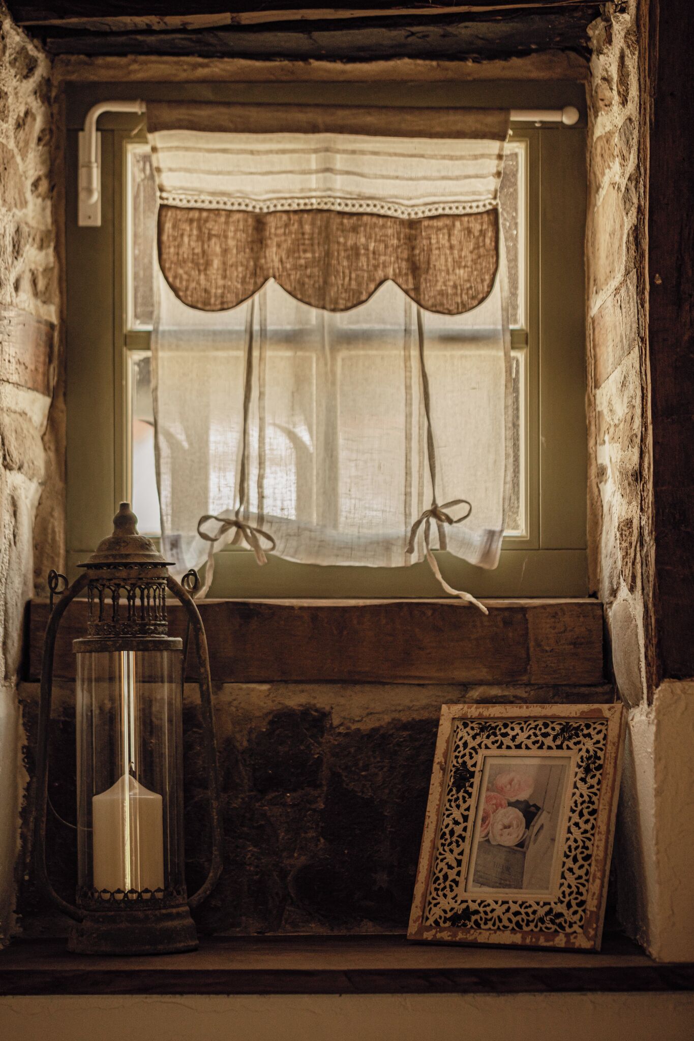 A rustic window with a curtain, a lantern, and a framed picture on a stone ledge.