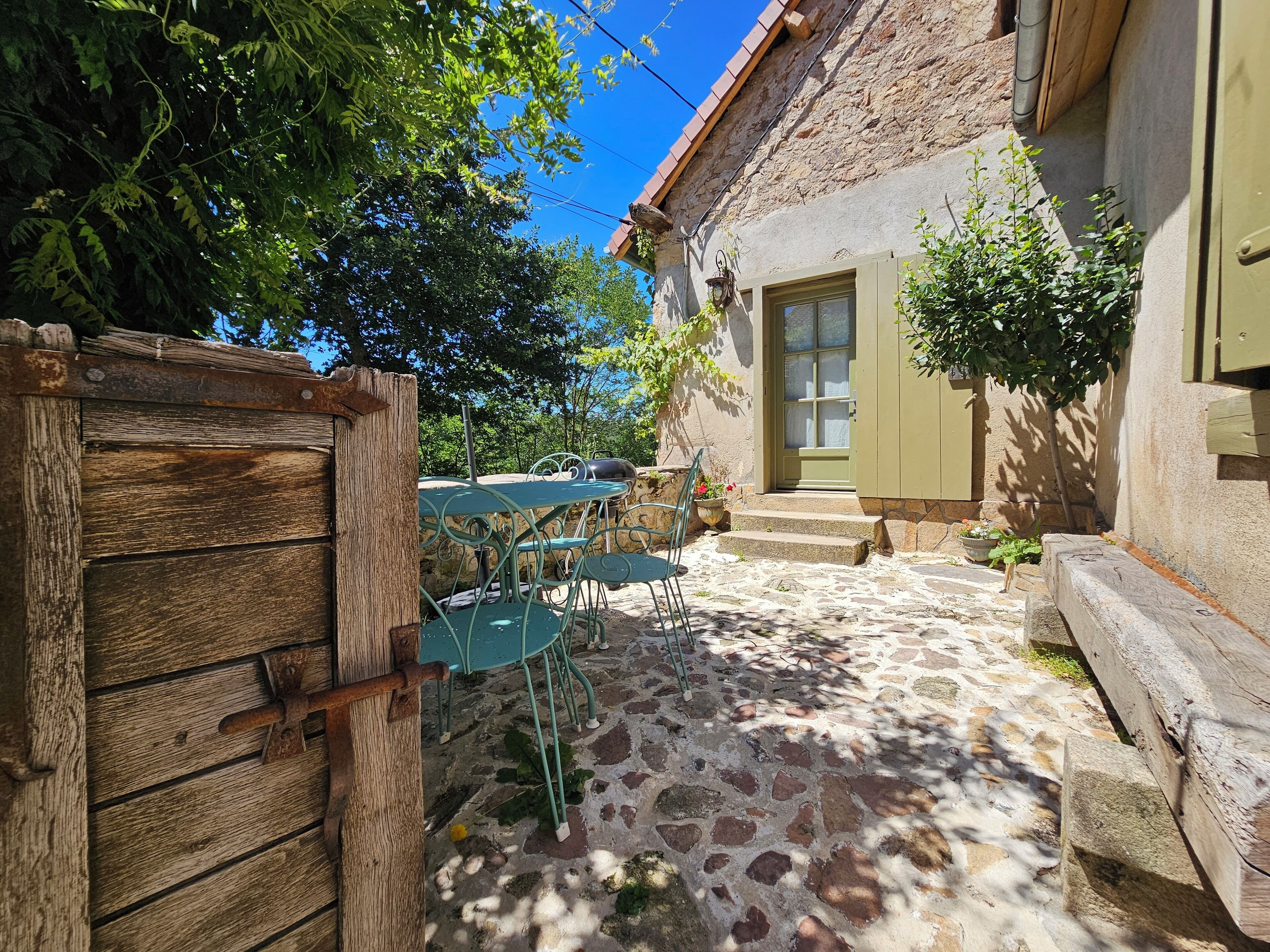 Une cour rustique en pierre, avec un portail en bois, une table en pierre entourée de chaises et un bâtiment doté d’une porte verte.