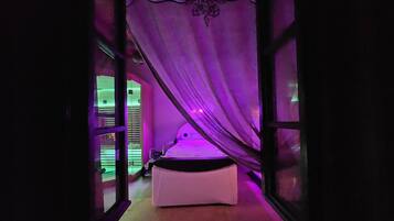 A room with a bed under a purple canopy, a window with blinds, and a door leading to another room.