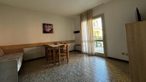 Una habitación pequeña y poco amueblada, con suelo de baldosas, una cama individual, una mesa con sillas, una ventana con cortinas y una puerta.