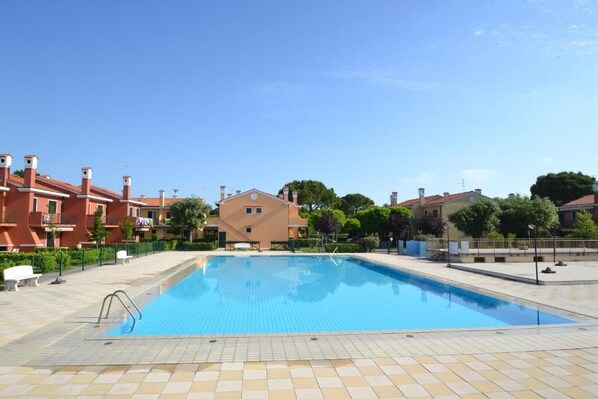 A swimming pool surrounded by buildings with red and beige facades, a clear blue sky, and some greenery.