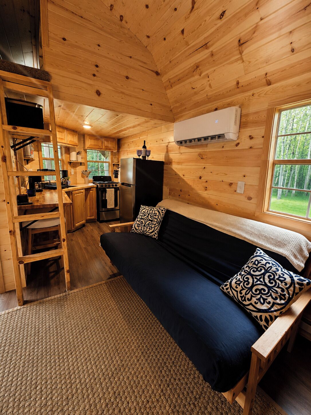 A cabin interior with a bed, kitchenette, and a window overlooking greenery.
