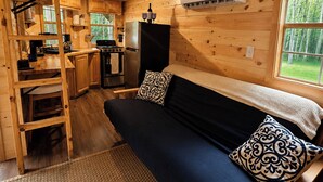 Interior de una cabaña con cama, kitchenette y ventana con vistas a la naturaleza.
