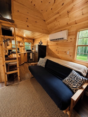 A cabin interior with a bed, kitchenette, and a window overlooking greenery.