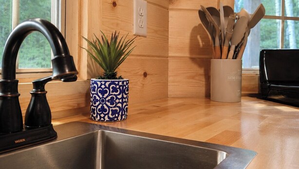 Una cocina con un lavamanos de acero inoxidable, armarios de madera, una planta en maceta y utensilios en un estante.