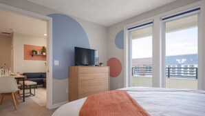 Una camera d'albergo con un letto, una televisione su un settimino in legno, un balcone con vista e un'area ristorazione con tavolo e sedie.