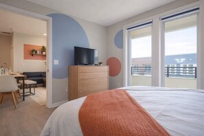 Habitación de hotel con cama, televisor en una cómoda de madera, balcón con vistas y zona de comedor con mesa y sillas.