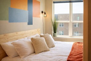 Quarto com cabeceira de madeira, roupa de cama branca e vista para um edifício de tijolos pela janela.