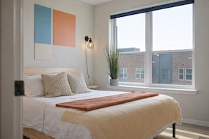 Un dormitorio con cama, almohadas, una planta en la mesita de noche y una ventana con vistas a edificios.