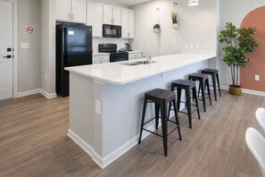 A modern kitchen with white cabinetry, a black refrigerator, a microwave, and a sink. There are four bar stools and a potted plant.