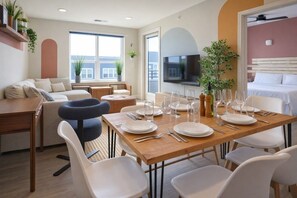 Un comedor con una mesa de madera preparada para seis personas, rodeada de sillas blancas. Se observa un sofá y un televisor al fondo.
