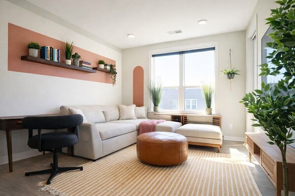 Una sala de estar moderna con un sofá, un reposapiés y un escritorio con una silla. Hay una ventana que ofrece vistas a edificios.
