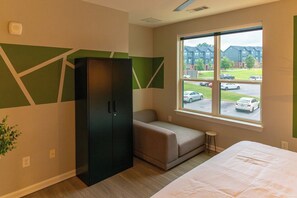 A modern hotel room with a bed, a seating area, a wardrobe, and a window with a view of the street.