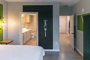 A bedroom with a bed, a small table, and a view into the bathroom and kitchen.
