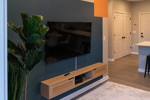 Een moderne woonkamer met een flatscreen televisie aan de muur, een houten tv-meubel eronder en een kamerplant aan de linkerkant.