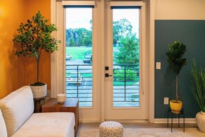 Habitación con sofá, una mesita de noche con lámpara, una planta en maceta y vistas a un aparcamiento a través de una puerta.