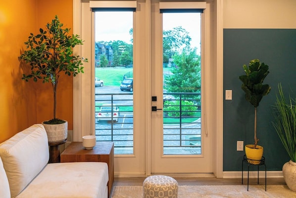 A room with a sofa, a small table with a lamp, a potted plant, and a view through a door to a parking area.