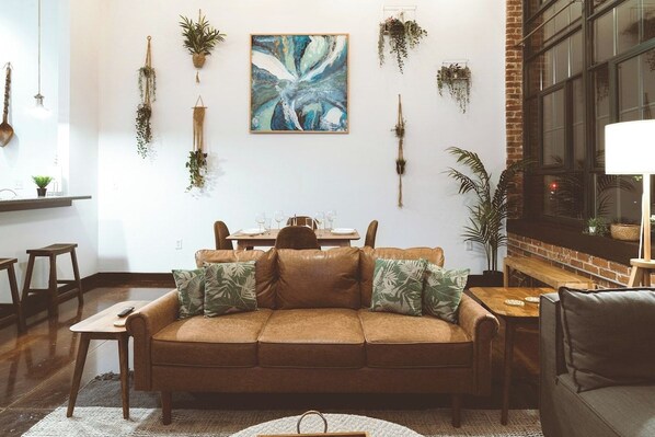 A cozy living room with a brown leather sofa, a dining table, and various plants on the walls. - Cozy Mill Loft w/ 2 king beds, huge windows (Spartanburg)