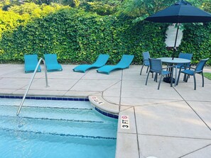 A swimming pool with lounge chairs and a table, surrounded by a hedge.