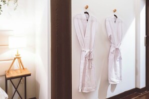 Dos batas blancas colgadas en una pared, una mesita de noche de madera con una lámpara y un suelo de madera oscura.