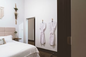 A hotel room with a bed, a nightstand, a wall-mounted macrame piece, and two white robes hanging on hooks.