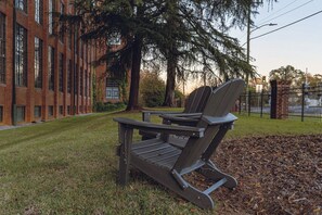 Un banco de parque con estructura metálica y listones de madera, situado en una zona cubierta de césped con un edificio de ladrillo al fondo.