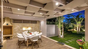 An outdoor dining area with a stone fireplace, a white table set for four, and a view of a well-maintained garden at dusk.