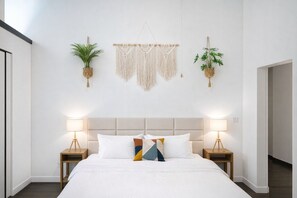 A bedroom with a bed, two bedside tables, a macrame wall hanging, and two hanging plants.